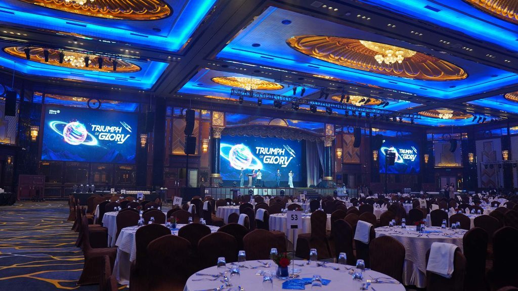 An elaborate setup for an international grand event, featuring a grand ballroom with blue and gold lighting, beautifully arranged round tables, and a large stage with LED screens displaying the event theme 'Triumph of Glory,' complemented by a high-end production and sound system.