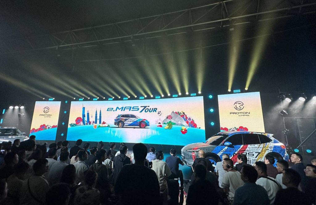 A car launch event showcasing the Proton e.MAS 7, featuring a vibrant panoramic LED screen stage backdrop with colorful graphics, including iconic landmarks and festive elements, complemented by dramatic lighting effects and an enthusiastic crowd in attendance.