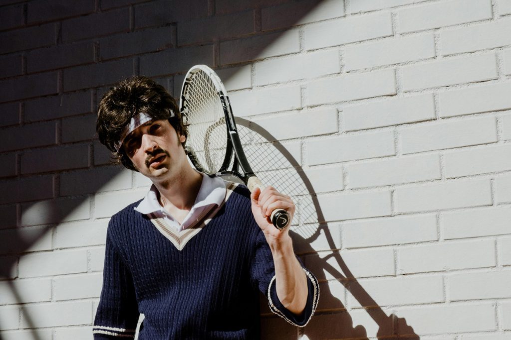 A person dressed in vintage tennis attire for a sports-themed annual dinner party. They are wearing a navy sweater with a white collar, a headband, and holding a tennis racket over their shoulder, evoking a classic retro sports look.